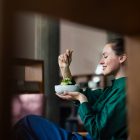 Emulsifiers and gut health - young woman eating healthy food in a bowl with a fork, in an office. She is smiling and the picture is taken from a distance