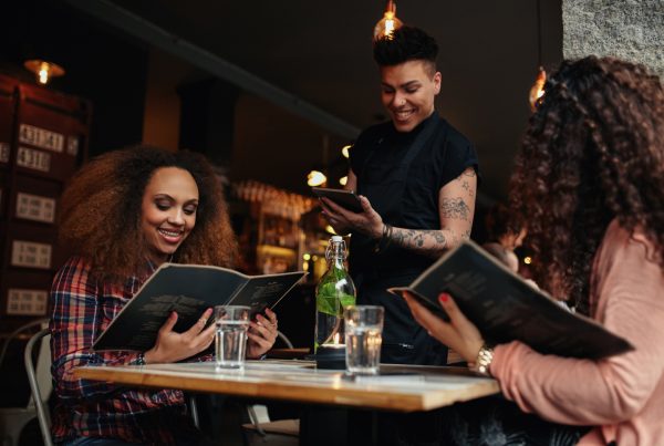 Young women seated at a table holding menus in a restaurant. Their waiter is standing waiting to take their order. Food combining part 2